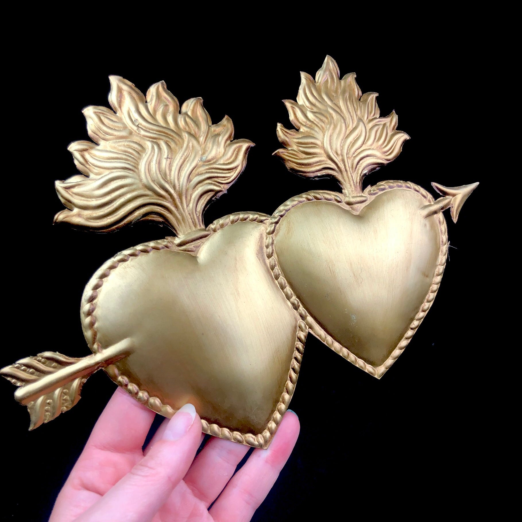 Two golden brass sacred hearts with flames held togther with an arrow that passes through them